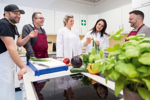 Einblick in eine Diät-Lehrküche mit drei männlichen Teilnehmern und zwei Diät-Assistentinnen, die die Zubereitung von Gemüse erklären.