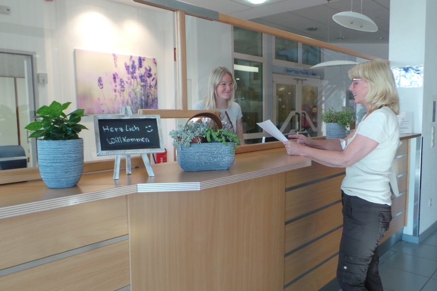 Eine freundlich lächelnde junge Frau steht hinter einem Empfangstresen. Davor steht eine andere Frau, die sich ein Formular durchliest. Eine freundlich lächelnde junge Frau steht hinter einem Empfangstresen. Davor steht eine andere Frau, die sich ein Formular durchliest.