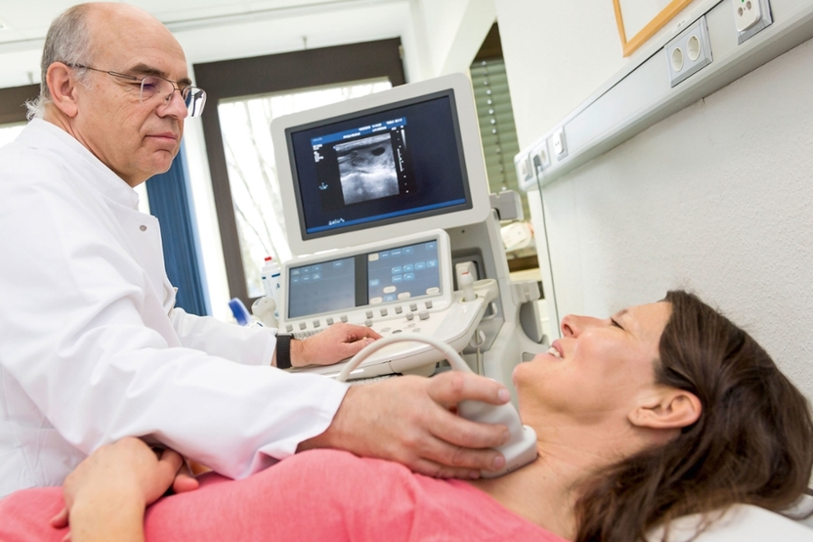 Ein Arzt nimmt bei einer Frau, die auf einer Behandlungsbank liegt, eine Sonographie an der Schilddrüse vor Ein Arzt nimmt bei einer Frau, die auf einer Behandlungsbank liegt, eine Sonographie an der Schilddrüse vor
