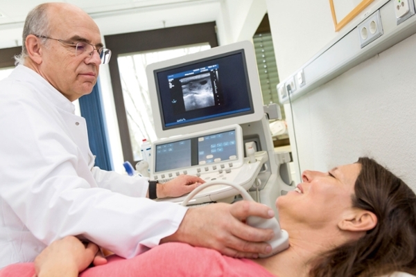 Ein Arzt nimmt bei einer Frau, die auf einer Behandlungsbank liegt, eine Sonographie an der Schilddrüse vor Ein Arzt nimmt bei einer Frau, die auf einer Behandlungsbank liegt, eine Sonographie an der Schilddrüse vor